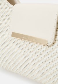 Tkana torebka w kolorze kremowym z gładką, usztywnioną klapką na górze. Złota plakietka z logo i fakturowane detale na powierzchni.