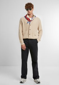 Cardigan bej cu dungi, cu decolteu în V, două buzunare frontale și nasturi maro. Asortat cu pantaloni negri și o eșarfă multicoloră.