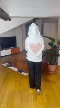 Sweatshirt branca com um grande coração rosa claro estampado nas costas, combinada com calças pretas; ambiente interior com plantas e mobiliário.