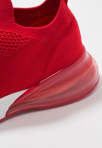 Červená sportovní bota s texturovaným pleteným svrškem a průhlednou vzduchovou podešví. Disponuje konturovaným designem a bílými akcenty.