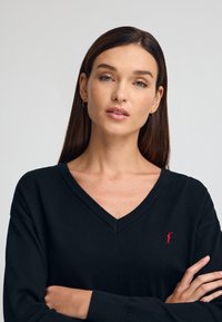 Czarny sweter w serek wykonany z dzianiny. Posiada długie rękawy oraz małe, czerwone haftowane logo po lewej stronie. Gładka tekstura.