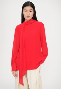 Blouse rouge à manches longues avec un nœud au col, réalisée en tissu léger. Comprend des poignets avec boutons et une coupe ample.
