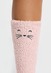 Calzino rosa, peloso, fino al ginocchio, realizzato in materiale morbido, con un volto di gatto ricamato in nero, complete di baffi e un'espressione sorridente.