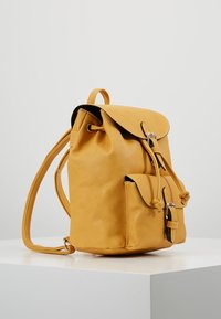 Hořčičně žlutý kožený batoh s přední kapsou, sponovými uzávěry a nastavitelnými ramenními popruhy na bílé ploše na jednobarevném pozadí.