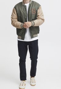 Groene en beige bomberjack met een opvallende "B" patch, geribbelde boorden en zoom, gedragen met donkere jeans en witte sneakers.