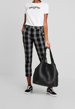 Personne portant un t-shirt blanc Hollister, un pantalon à carreaux noir et blanc, des baskets noires, tenant un grand sac fourre-tout noir tissé.