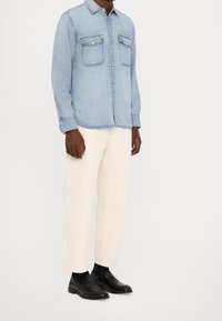 Camisa de mezclilla azul claro con botones, dos bolsillos en el pecho, combinada con pantalones color crema y mocasines negros, con un ajuste relajado y mangas largas.