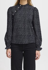 Blusa nera con pois bianchi, scollo alto, dettaglio arricciato con bottoni decorativi, maniche lunghe con polsini arricciati, tessuto morbido.