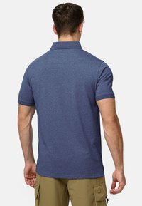 Man draagt een polo van gemêleerd blauw met korte mouwen en olijfgroene cargoshorts, staand met de rug naar voren tegen een witte achtergrond.