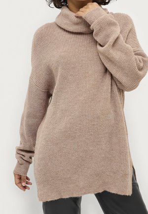 Femme portant un pull à col roulé en maille beige oversize avec manches longues et fentes sur les côtés, associé à un pantalon noir.
