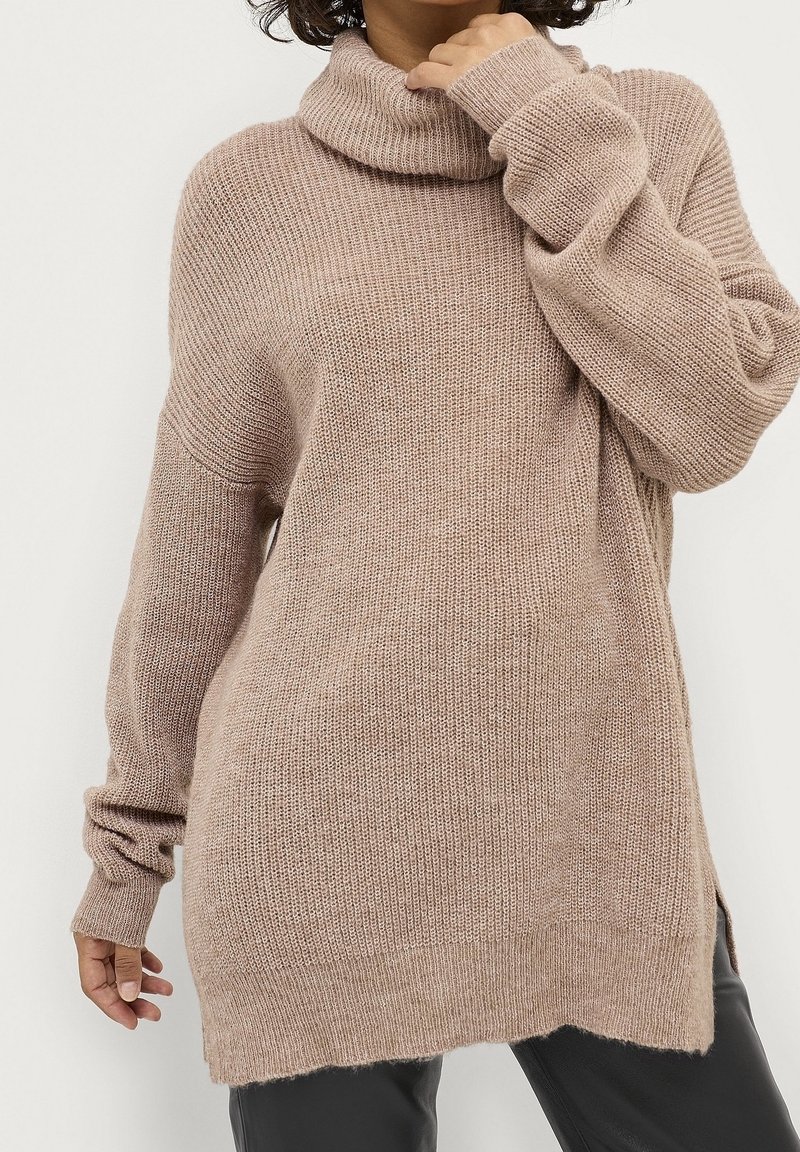 Femme portant un pull à col roulé en maille beige oversize avec manches longues et fentes sur les côtés, associé à un pantalon noir.