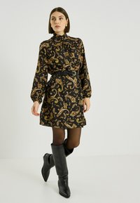 Vestido negro con patrón de cachemira dorado y verde, cuello alto, mangas largas y cintura ceñida con cinturón. Combinado con botas negras hasta la rodilla y medias transparentes.