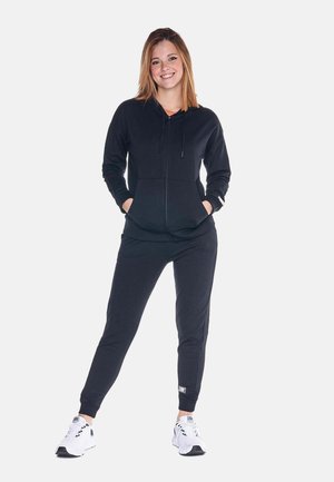 Une femme souriante porte un sweat à capuche noir zippé assorti à un pantalon de jogging, avec des baskets blanches, sur un fond blanc uni.