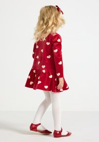 Robe en velours rouge avec des motifs de cœur rose, ourlet volanté, collants blancs et chaussures rouges brillantes. Cheveux coiffés avec un accessoire en forme de nœud rouge.