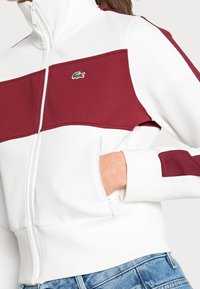 Person som bär en vit jacka med vinröda paneler och krokodil-logga, med handen i dragkedjefickan, tillsammans med blå denimjeans.