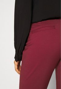 Pantalon bordeaux au toucher lisse avec une poche arrière, associé à un blouse noire à manches longues avec des poignets boutonnés.