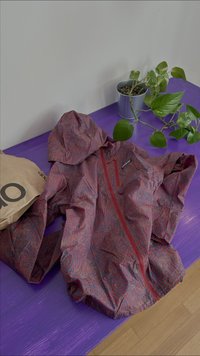 Rood en blauw patroon lichtgewicht jack met capuchon, rits aan de voorkant en lange mouwen, geplaatst op een paarse ondergrond naast een plant en een tas.