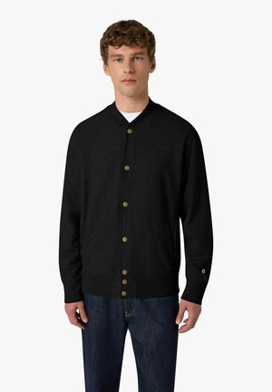 Giovane uomo che indossa un cardigan nero con bottoni dorati, maglietta bianca sotto e jeans blu, in piedi davanti a uno sfondo semplice.