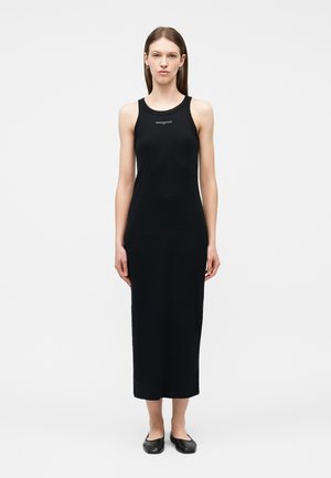 Žena stojaca, oblečená v čiernych midi šatách bez rukávov s malým bielym nápisom na hrudi a čiernych rovných topánkach, neutrálne pozadie.