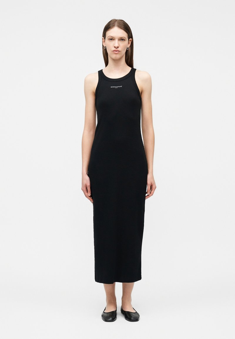 Γυναίκα που στέκεται φορώντας μαύρο midi φόρεμα χωρίς μανίκια με μικρό λευκό κείμενο στο στήθος και μαύρα φλατ παπούτσια, ουδέτερο φόντο.