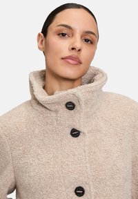 Beżowa polarowa kurtka z wysokim kołnierzem, z czarnymi guzikami i textured powierzchnią. Skupia się na ciepłe i komforcie w designie.