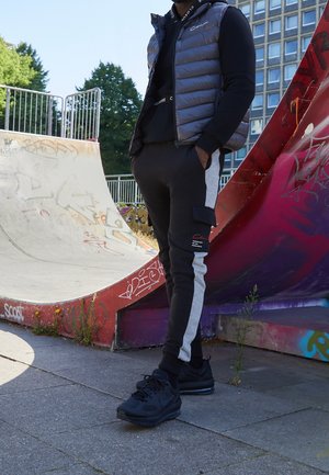 Vadderad grå väst över svart hoodie, svarta joggers med grå sidostripes och cargo-fickor, i kombination med svarta sneakers vid en skatepark.