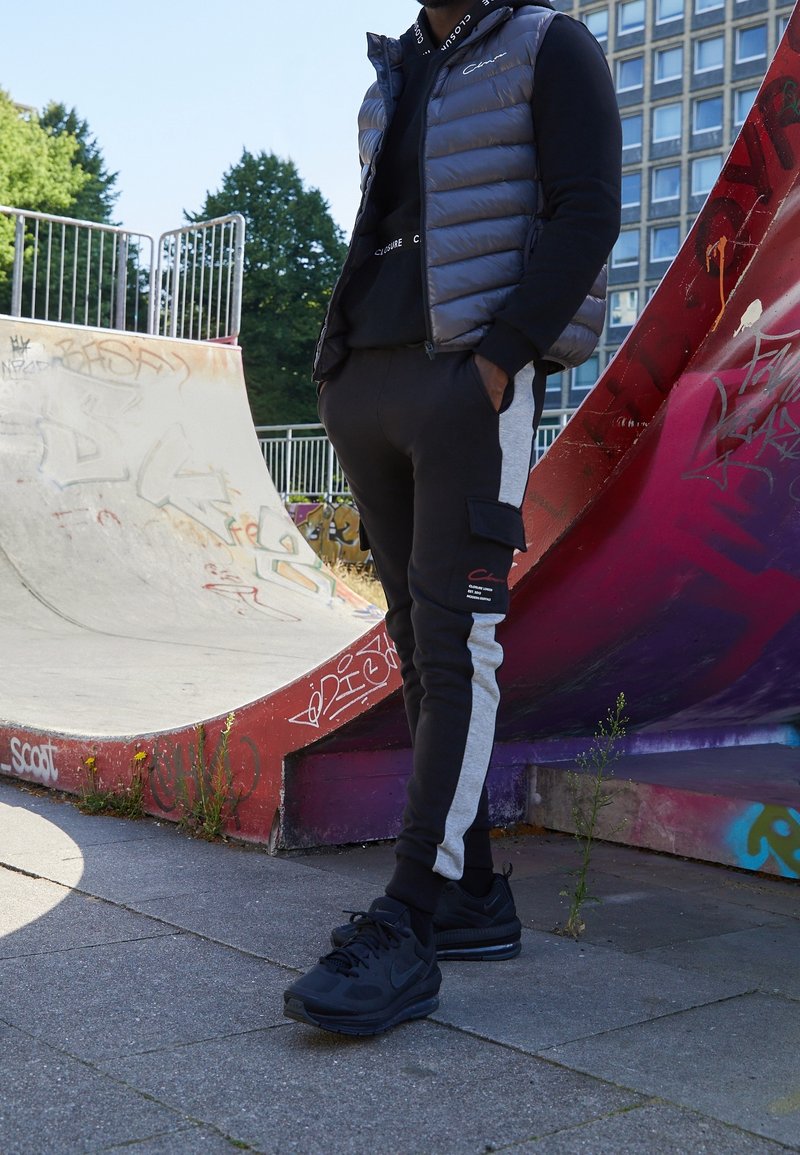Pěnová šedá vesta přes černou mikinu s kapucí, černé tepláky se šedými bočními pruhy a kapsami na cargo, doplněné černými teniskami ve skateparku.