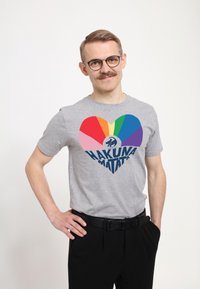 Šedé tričko s krátkým rukávem s velkým barevným srdcem a textem "HAKUNA MATATA" v námořnické modři. Nošeno s černými kalhotami.