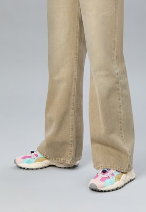 Persoon die beige broek met wijde pijpen en pastelkleurige sneakers draagt, staand op een effen grijze ondergrond.