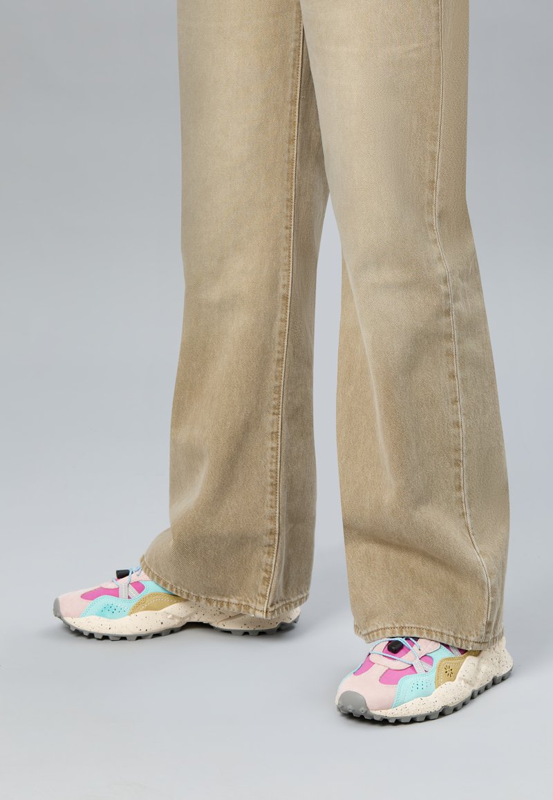 Person, die beige weite Hosen und pastellfarbene Turnschuhe trägt, steht auf einer schlichten grauen Fläche.