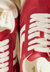 Raudoni odos ir zomšos sportiniai bateliai su baltais akcentais, turi plokščias smėlio spalvos raištelius ir tekstūruotą vidinę pamušalą. Ant vidpadžio išspaustas tekstas.