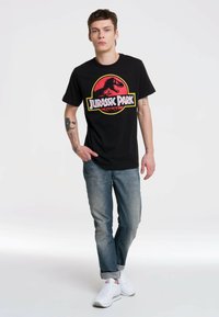 Camiseta de algodón negra con un gráfico del logo de Jurassic Park en rojo y amarillo. Combinada con jeans azul claro y zapatillas blancas.