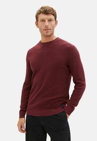 Bordeauxfarbener Rippenstrickpullover mit langen Ärmeln und Rundhalsausschnitt. Verfügt über ein dezentes Markenzeichen am Saum und hat einen regulären Schnitt.