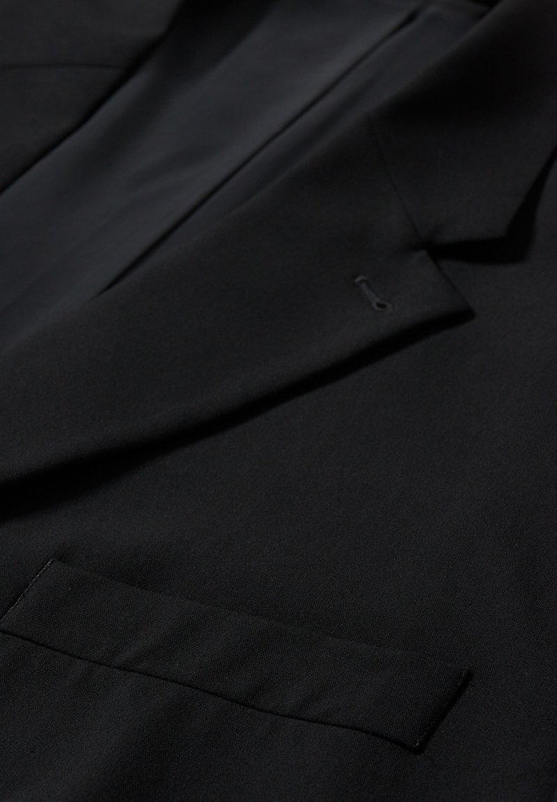 Detalle de la manga y el solapa de una chaqueta de traje negra, mostrando la textura de la tela, un ojal y un bolsillo en el pecho con costuras limpias.