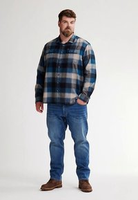 Camicia in flanella a quadri blu e beige con due tasche sul petto, chiusura con bottoni e maniche lunghe; indossata con jeans blu medio e stivali marroni.