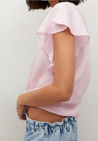 Donna che indossa una blusa senza maniche di raso rosa chiaro con volant, infilata in jeans di denim carta da zucchero blu chiaro a vita alta, su uno sfondo semplice.