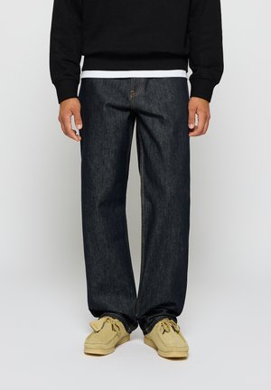 Dark blue denim jeans with a relaxed fit, featuring yellow stitching, a front pocket, and worn with beige moccasin-style shoes.