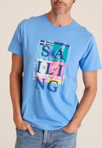 Lichtblauwe katoenen t-shirt met een kleurrafiek met zeilmotieven en tekst, ronde halslijn en korte mouwen.