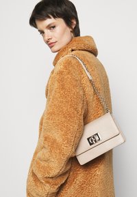 Borsa a tracolla in pelle beige con hardware dorato, forma rettangolare e finitura testurizzata, indossata sopra un cappotto in pelliccia sintetica marrone.