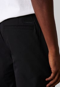 Pantalones negros con un bolsillo trasero que presenta una etiqueta texturizada de Calvin Klein. Tejido suave con un corte entallado, sin patrones ni acentos visibles.