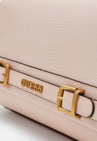 Détail d'un sac à main texturé rose clair avec boucle dorée et nom de la marque GUESS en lettres dorées sur une sangle.