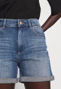 Jeansshorts med uppvikta fållar, bärs tillsammans med en svart topp, visar framfickor och knappstängning på en mörkhyad person.