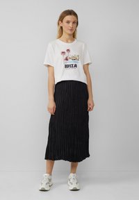 T-shirt blanc avec un graphisme représentant des palmiers et le texte "IBIZA BALEARIC PARADISE", associé à une jupe midi plissée noire et des baskets.