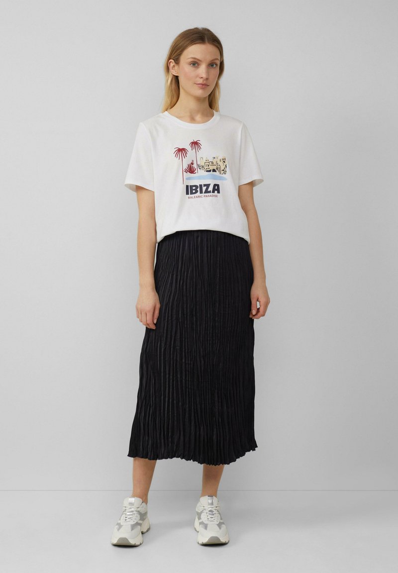 T-shirt blanc avec un graphisme représentant des palmiers et le texte "IBIZA BALEARIC PARADISE", associé à une jupe midi plissée noire et des baskets.