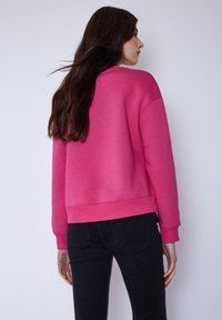 Rosa Sweatshirt mit lockerer Passform, gerippten Bündchen und Saum. Glatte Textur und mittleres Gewicht, kombiniert mit schwarzer Jeans. Rückansicht gezeigt.