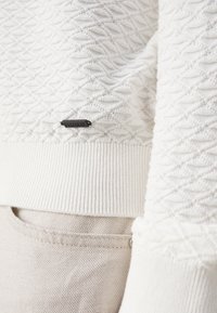 Maglione bianco con texture, orli e polsini a coste; presenta un piccolo etichetta nera con la scritta "HUGO". Sullo sfondo si trova un tessuto beige chiaro.