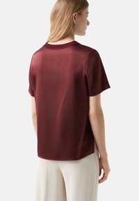 Bordeaux kortærmet top lavet af glat stof med en rund halsudskæring og en lidt længere bagkant. Enkelt design, ingen mønstre eller detaljer.