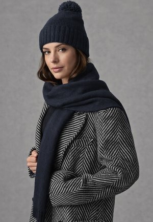 Frau trägt eine dunkle gestrickte Mütze mit Bommel, einen passenden Schal und einen Mantel mit Fischgrätmuster vor einem einfarbigen grauen Hintergrund.