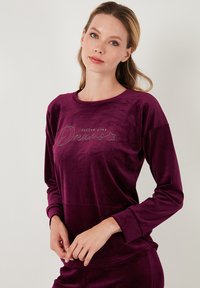 Long-sleeve, deep purple velour top with "follow your Dreams" text in metallic green. Round neckline and fitted sleeves. Soft texture.