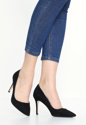 Pieds portant des escarpins en daim noir associés à un jean skinny bleu raccourci au-dessus des chevilles sur fond blanc.